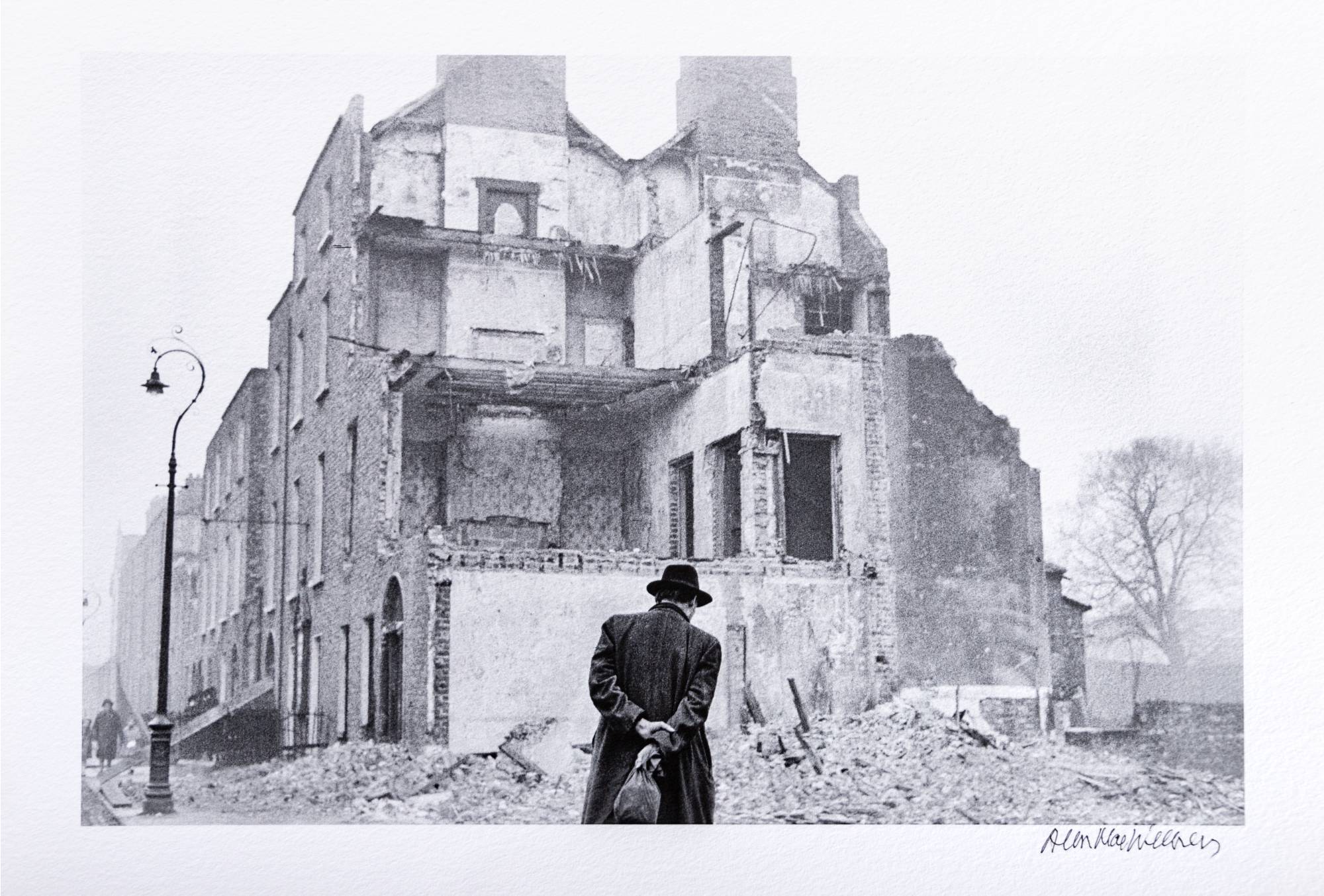 man in long dark coat looking at the remains of a partially destroyed residential building